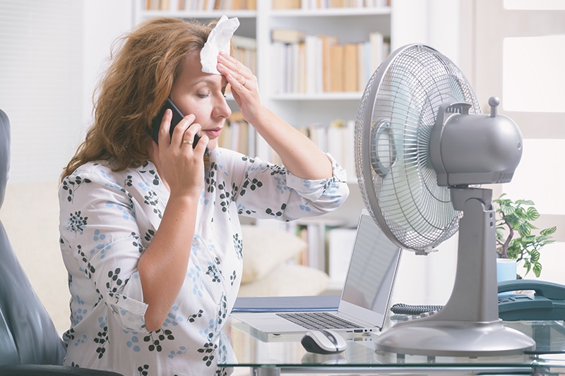 Woman sitting in front of a desk fan wiping her damp face due to a failed air conditioner.