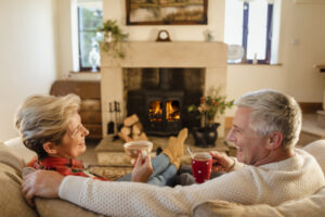 Older couple sitting in front of a fire drinking hot chocolate from mugs.