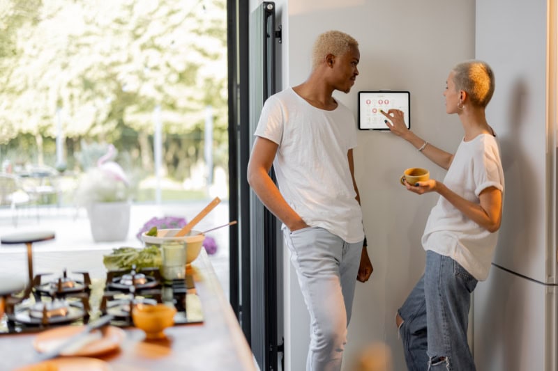 Girl choosing temperature on thermostat of smart home system while her boyfriend looks at her.